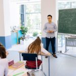 Teacher instructing students in a bright classroom with a blackboard filled with math equations.